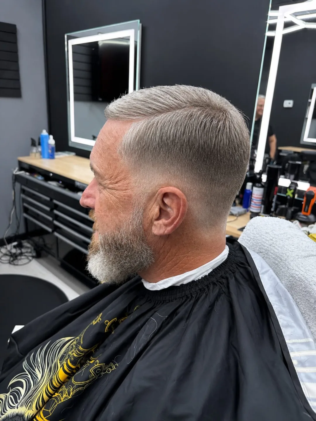 Man getting a faded haircut in a barbershop, seated in a cape beside a mirror and grooming tools.
