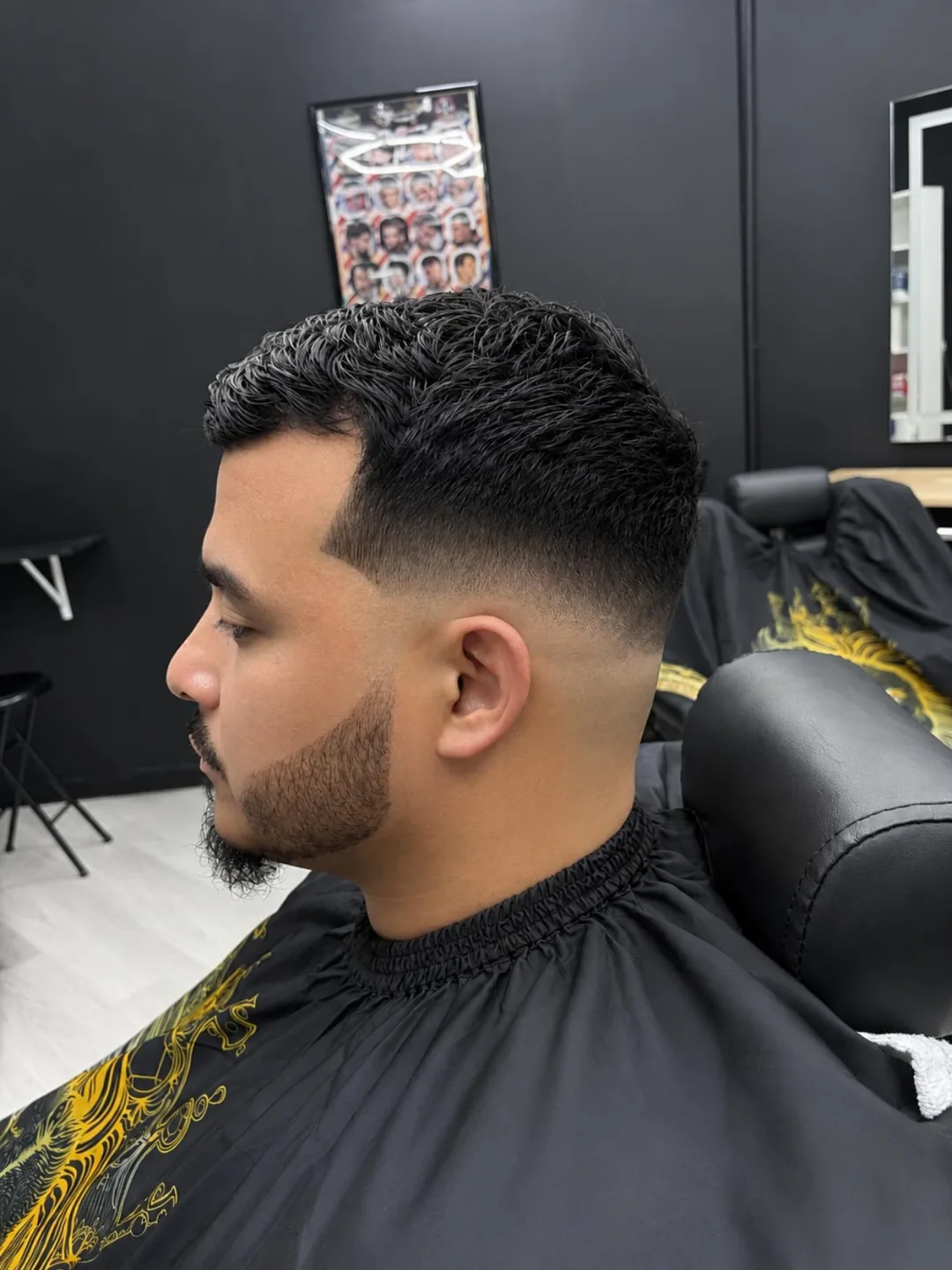 Side-profile haircut in a barbershop, with a fade and curly top, draped in a black cape.